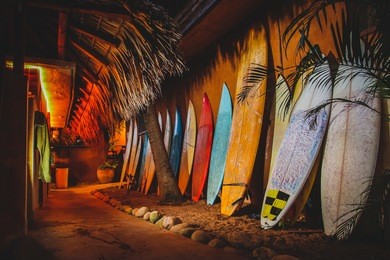 a collection of different colorful surf boards used as a decoration next to a walkway, resting on a wall. photo taken during the night with a bunch of surfboards on the sand.