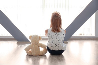 little girl with teddy bear sitting on floor near window in empty room. autism concept
