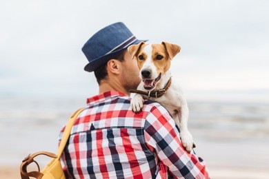 small beautiful dog on owner`s hands. stylish young male model dressed in fashionable clothes carries his pet and bag, stand at seashore, come to admire wonderful landscapes and breath fresh air