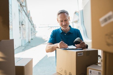 delivery man signing checklist and holding scanner in hand. happy courier writing on clipboard and scanning barcode. mature man updating checklist of delivery in van.