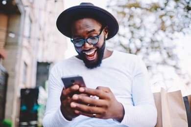 emotional surprised african american guy with low prices in web store receiving message with promo code,excited dark skinned hipster guy in trendy hat overjoyed with winning online contest on web site