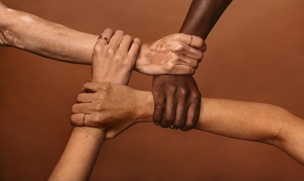 four diverse women holding each others wrists in a circle. top view of female hands linked in the lock against brown background.
