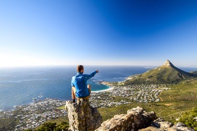 young male hiker sitting on a rock at kasteelspoort hiking trail in table mountain national park, cape town, south africa, pointing at the suburb of camps bay and lion`s head mountain in background.