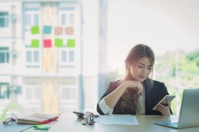 young asian business woman using laptop computer and cell phone for contact customer on wooden desk in office. business portrait concept.