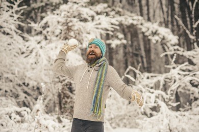 snow fight, sport, rest. bearded man smile with snowballs in snowy forest. temperature, freezing, cold snap. skincare, beard care in winter. hipster in thermal jacket, hat, scarf, beard warm in winter