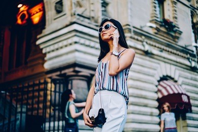 serious female in sunglasses and trendy outfit talking on phone walking on city street. attractive young woman having mobile conversation using connection in roaming strolling during free time