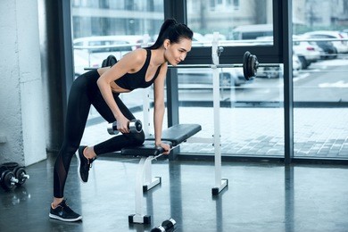young sporty woman lifting dumbbell while leaning on workout bench at gym