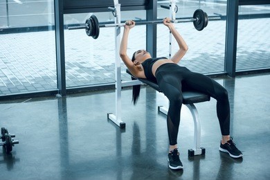 young athletic woman lifting barbell while lying on bench at gym