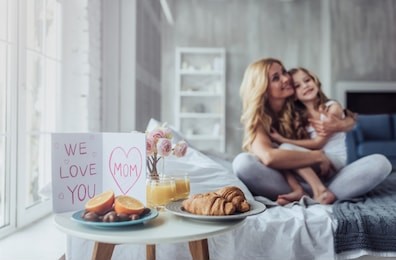 attractive young woman with little cute girl are spending time together at home while sitting on bed. happy family concept. breakfast in bed on mother's day.