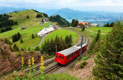 a sightseeing train traveling on the cogwheel railway through green grassy meadows on mt. rigi, with rugged pilatus peaks among majestic mountains in background on a cloudy summer day in switzerland