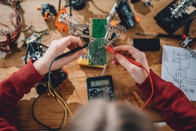 girl checking circuit board with multimeter on the wooden table at home and building a robot as a school science project