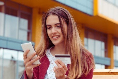 cheerful young pretty woman using smartphone and credit card for payment on the street. 