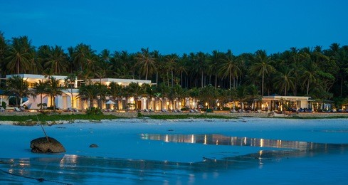 dusk at patok beach (racha island, phuket, thailand); the tide is low, the buildings are lit up and in the background the coconut trees sway in the wind. 