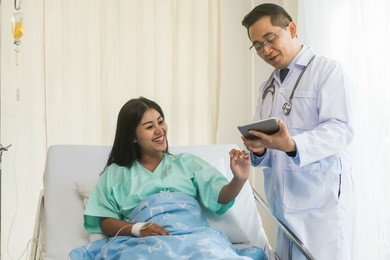 cheerful asian doctor talking with young woman patient for monitoring and check up after surgery. doctor giving advice in the touchscreen to women to take care the lesion after out of hospital.