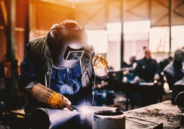 profesional welder in protective uniform and mask welding metal pipe on the industrial table with other workers behind in the industrial workshop.
