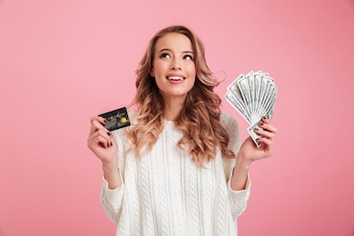 photo of happy young woman standing isolated over pink background. looking aside holding money and credit card.