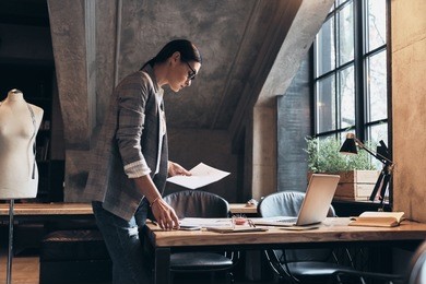 is it good enough? concentrated young woman in eyewear looking at sketches while leaning on the desk in her workshop 