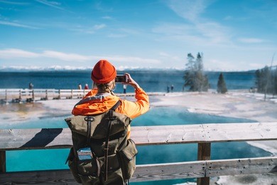 back view of female tourist with backpack using smartphone for taking picture of hot springs, hipster wanderlust making image on smartphone camera of touristic place in usa with geyser in yellowstone