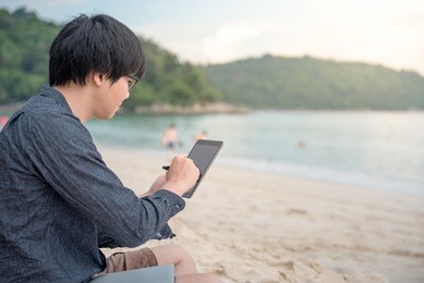 young asian man using tablet on tropical beach, digital nomad or freelance lifestyle concepts