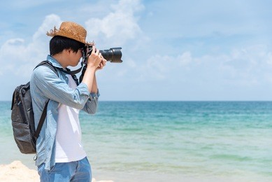 young asian man photographer with jean shirt and hat take photo of tropical island beach and turquoise sea, seascape background for summer holiday and vacation travel concepts