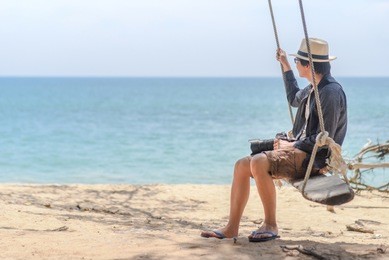 young asian photographer man on wooden swing at the beach, male traveler relaxing with seascape in summer season of thailand, travel lifestyle concept