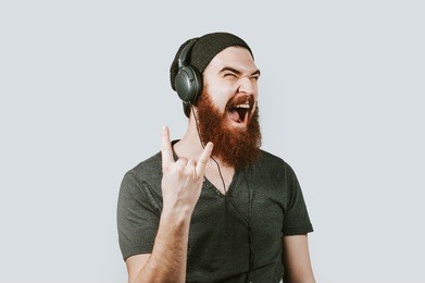 cheerful happy bearded hipster man listen music at headphones and show rock and roll sign. man screaming and listening music over grey background.