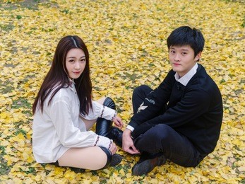 portrait of couple of chinese young people sit on the ground covered with yellow leaves in park face to face, and looking to camera together, lover concept.