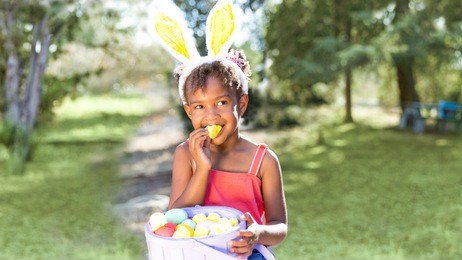 cute and beautiful african american girl wearing bunny ears, eats with joy easter marshmallow candy while holding easter basket full of eggs. girl sits in park outdoors in spring. text copy space.