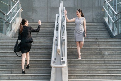 two business asian lady wave their hands to each other when they walking on the stairs.they both wearing a big smile.on a side view. wearing formal suits and skirts. 