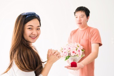 happy beautiful asian woman looking at camera while man propose to marry by giving red ring box and flower bouquet  isolated on white with copy space for text. 2018 valentine and wedding  celebration.