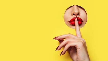 red lips look through the hole in the yellow paper. a finger with a red varnish at the mouth.