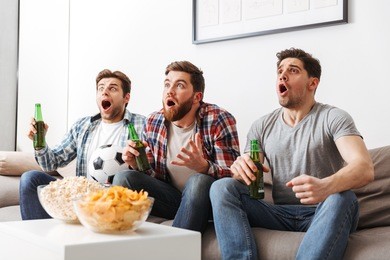 portrait of three excited young men watching football while sitting at home, drinking beer and eating snacks