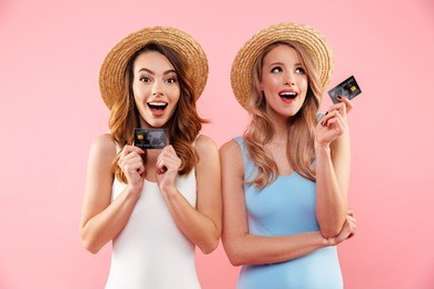 portrait of a two satisfied girls dressed in swimsuits and summer hats holding credit cards isolated over pink background