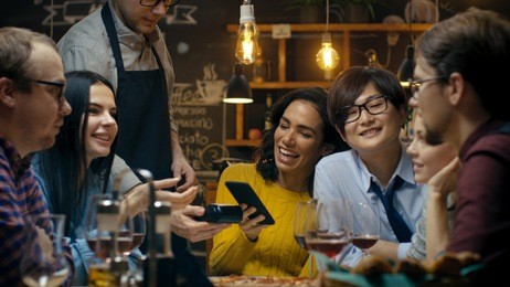 in the bar waiter holds credit card payment machine and beautiful woman pays for her order with contactless mobile phone payments system. she has good time with her friends.