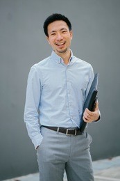 portrait of a young, chinese, pan asian business man standing against a plain grey background and smiling while he holds his leather folio. he is well-dressed in business casual and looks relaxed. 
