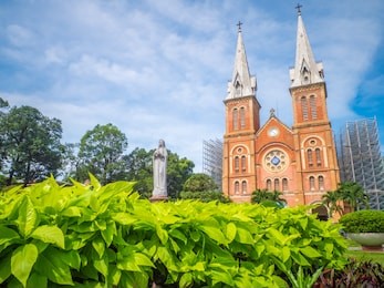 a famous christian church and maria statue with foreground green plant and blue sky. name is notre-dame cathedral basilica of saigon in ho chi minh city, vietnam.