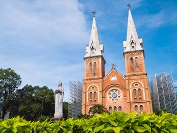 a famous christian church and maria statue with foreground green plant and blue sky. name is notre-dame cathedral basilica of saigon in ho chi minh city, vietnam.