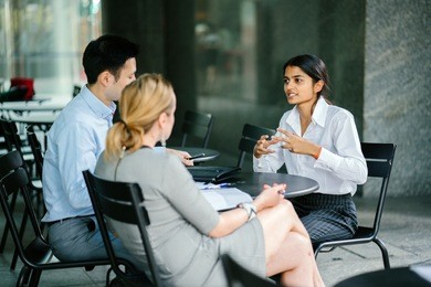 a young indian woman is interviewing for a job and speaking to a diverse panel of interviewers -- one is a chinese man, the other a caucasian white woman. they are sitting and speaking animatedly.