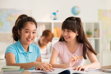 cheerful teenage students having fun while sitting at desk and doing assignment together, interior of modern classroom on background
