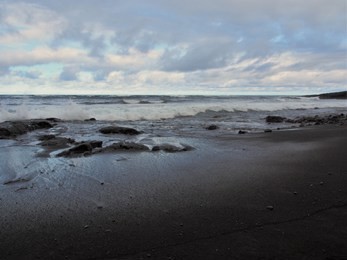 punalu'u black sand beach with shining wet sand, distant calm waves, rocks, and cloudy sky, the big island, hawaii
