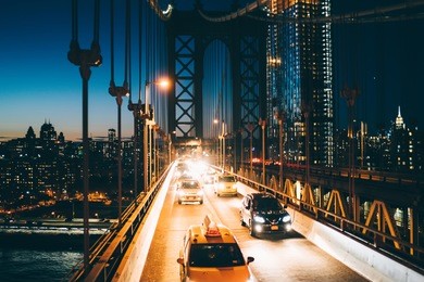 metropolitan traffic on brooklyn bridge with vehicles shining with evening light, yellow cab taxis driving from manhattan to another district, river crossings, environmental impact reduction concept