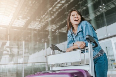 happy young attractive asian woman passenger with luggage trolley walking through an airport