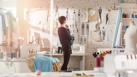 female fashion, designer,  looking at drawings and sketches that are pinned to the wall behind her desk. studio is sunny. personal computer, colorful fabrics, sewing items are visible.