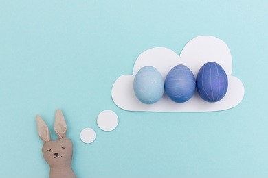 plush easter bunny with closed eyes and dream cloud with blue colored easter eggs. funny easter preparation concept as invitation for egg painting easter party.