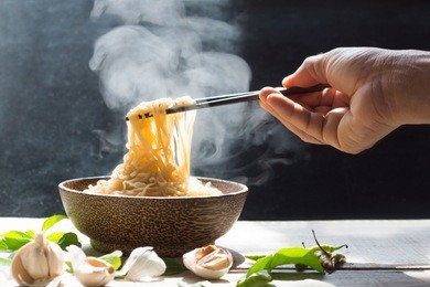 hand uses chopsticks to pickup tasty noodles with steam and smoke in bowl on wooden background, selective focus. asian meal on a table, junk food concept