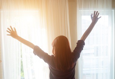 
rear view of a woman in plaid shirt standing near the window while stretching near bed after waking up with sunrise at morning,beautiful woman looking outside the window in her beautiful apartment,