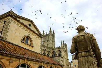 bath (england, uk) stone statue of the roman in antique roman baths complex, flying birds in sky and abbey cathedral at background. city of bath is a unesco world heritage site. 
