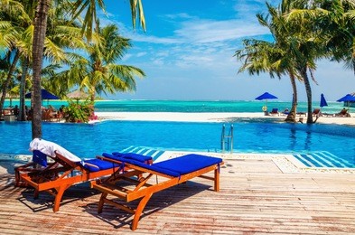 wooden deck chairs with blue pillows above the pool. in the background, the azure water of the ocean and a sandy beach with palm trees.