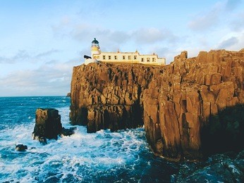 neist point lighthouse on rocky cliff above wavy sea. blue evening sea and sharp cliffs, isle of skye, scotland