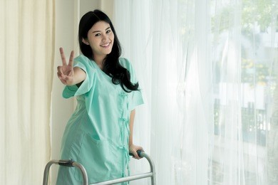 young asian patient woman standing with folding walker at hospital room showing victory sign for cheerful.